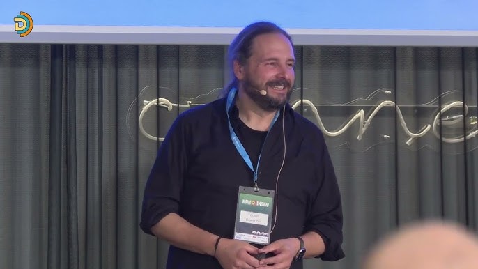 Tobias Goeschel speaking at KanDDDinsky 2024. He smiles and wears a headset microphone and a KanDDDinsky badge and lanyard.
